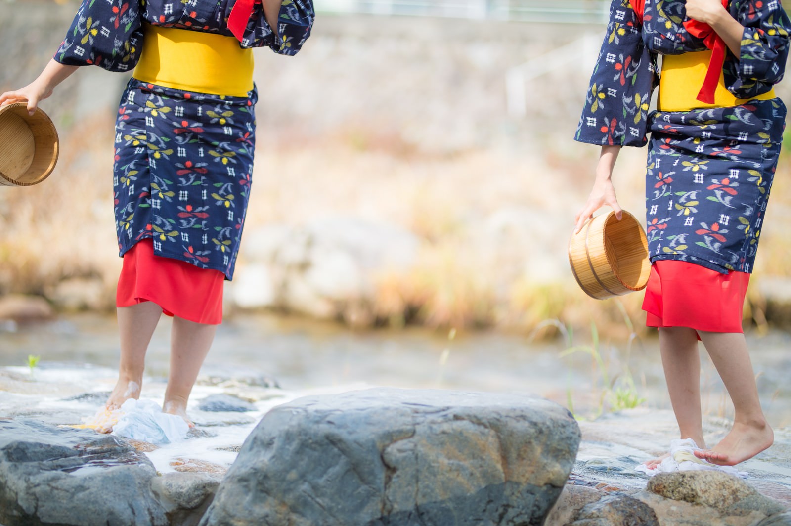 Women in yukata holding wooden buckets practicing traditional foot-stomping laundry on rocks