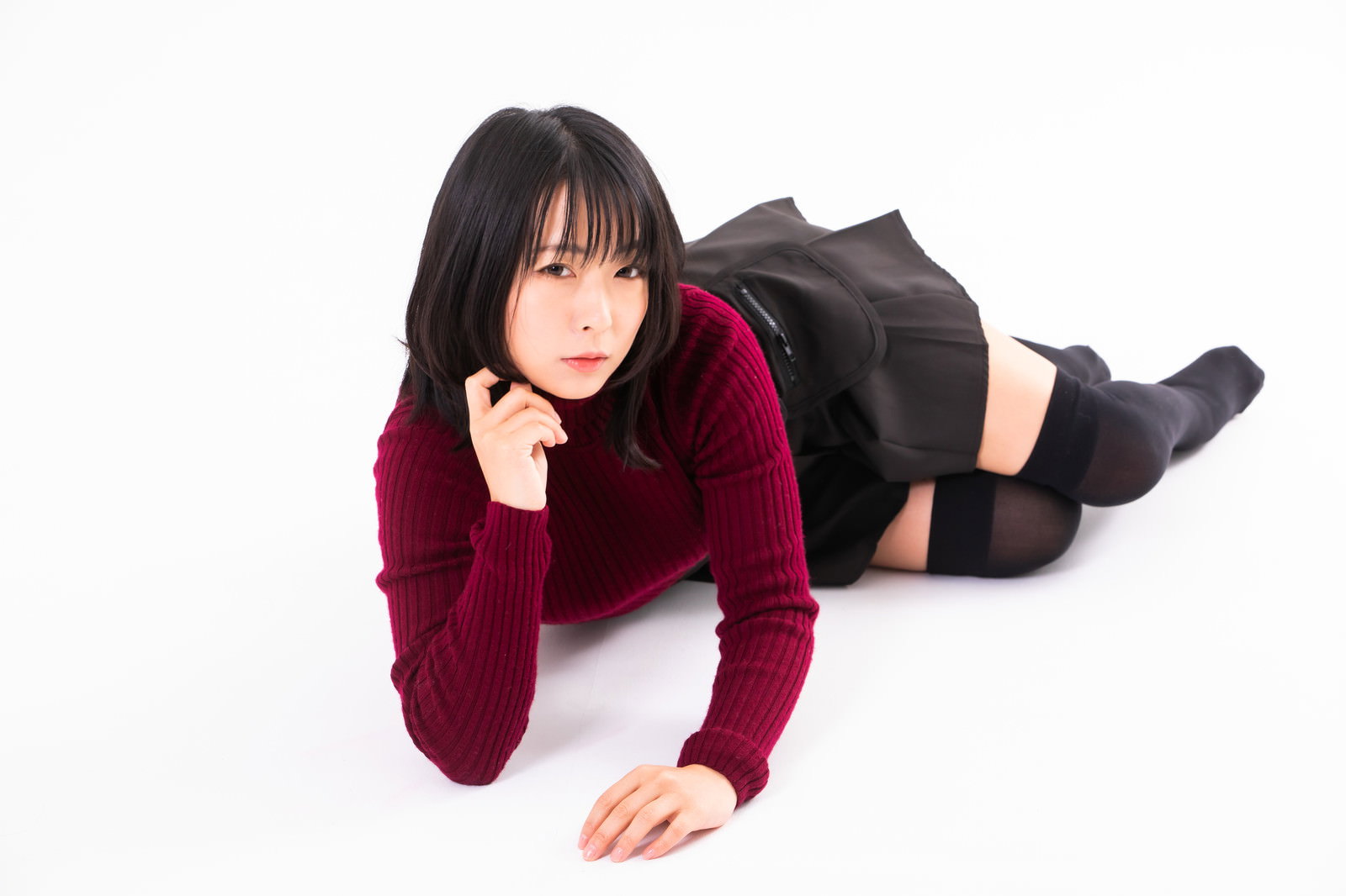 エンジ色のニットを着て横になり、肘をついてこちらを見つめる女性［モデル：にゃるる］