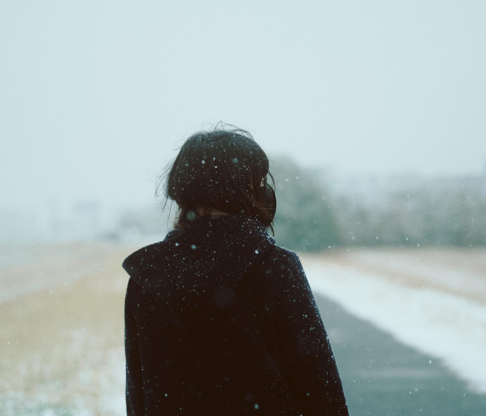 粉雪が降る冬の道で、黒いコートを着た女性が後ろ向きに立っている