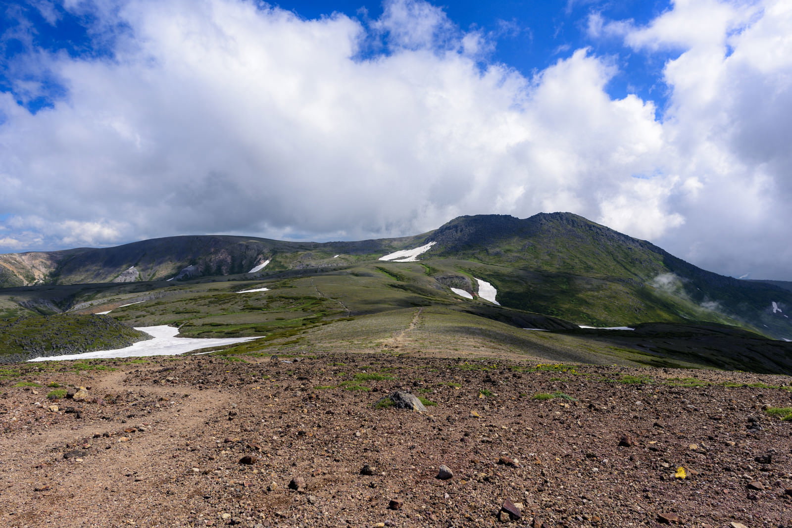青空と雲の下、緑の山々が連なる稜線を歩く登山道