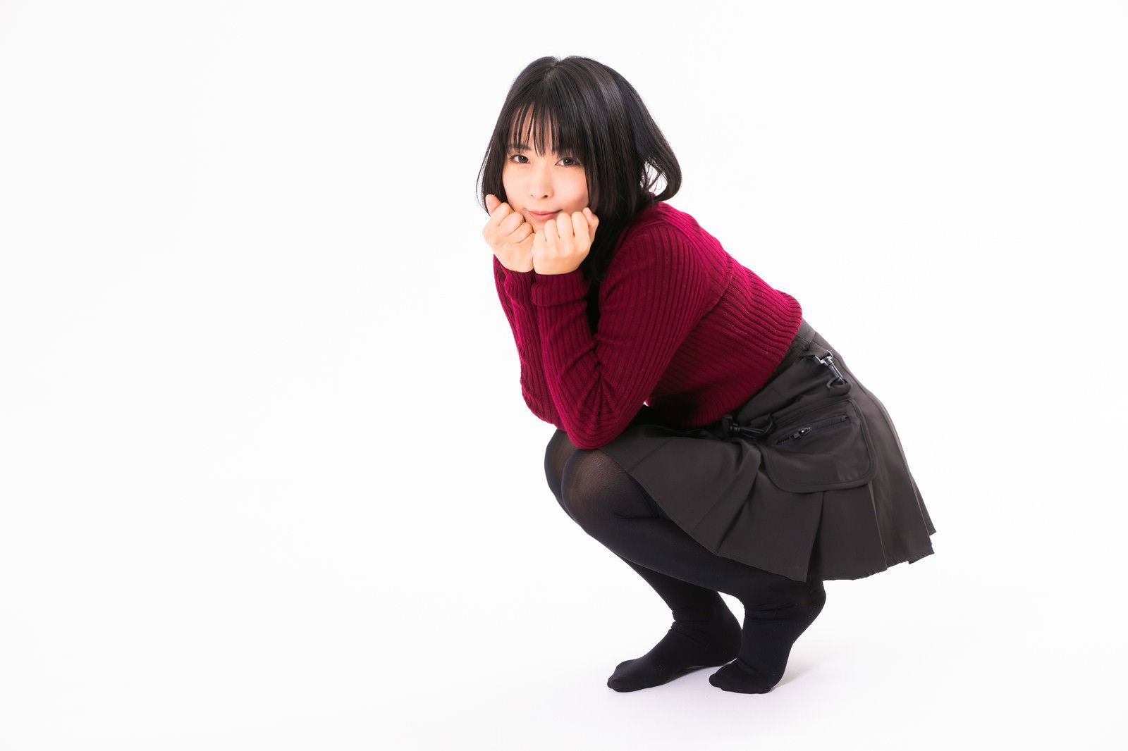 赤いニットを着た女性が照れながらしゃがみ込んでいる［モデル：にゃるる］