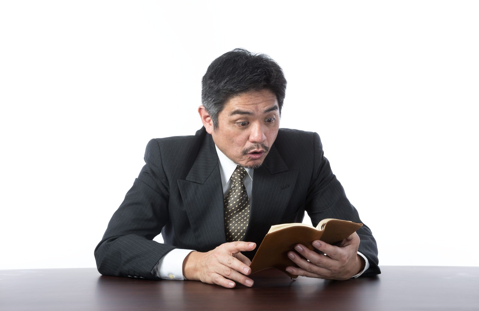 黒いスーツの男性がデスクで手帳を開き、驚いた表情で上を見ている［モデル：よたか］