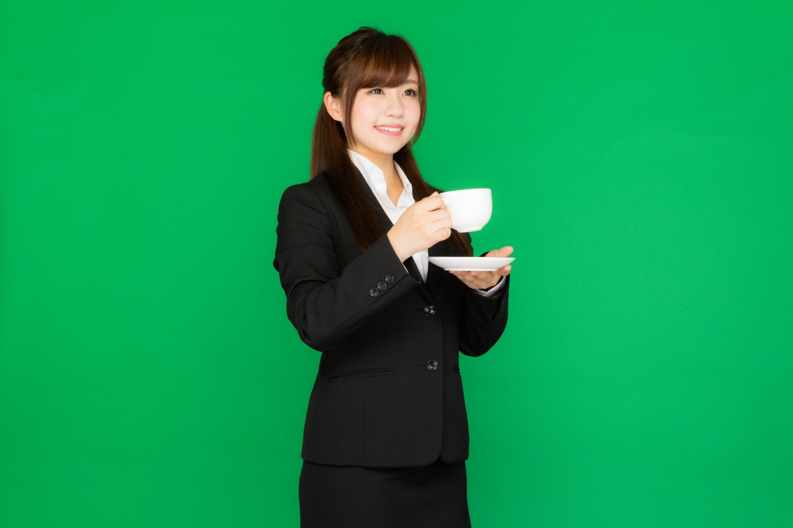 グリーンバックの前でコーヒーカップを両手で持つスーツ姿の女性［モデル：河村友歌］