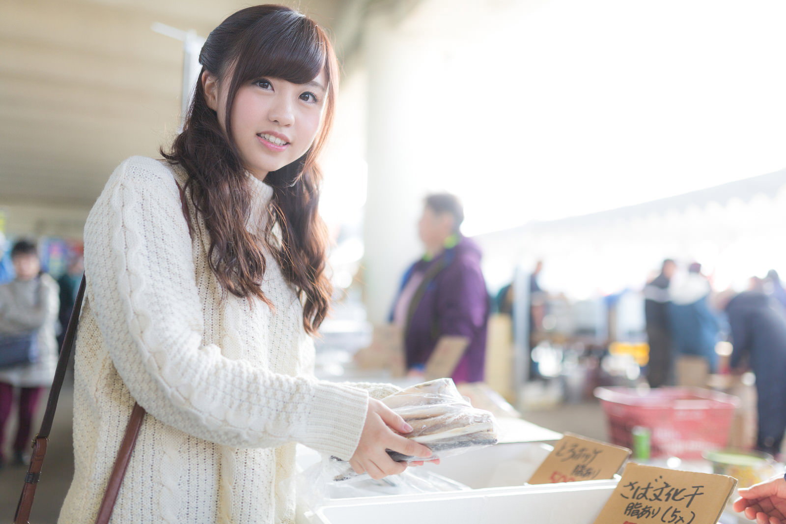 朝市の野菜コーナーで品物を手に取る白いシャツ姿の若い女性［モデル：河村友歌］