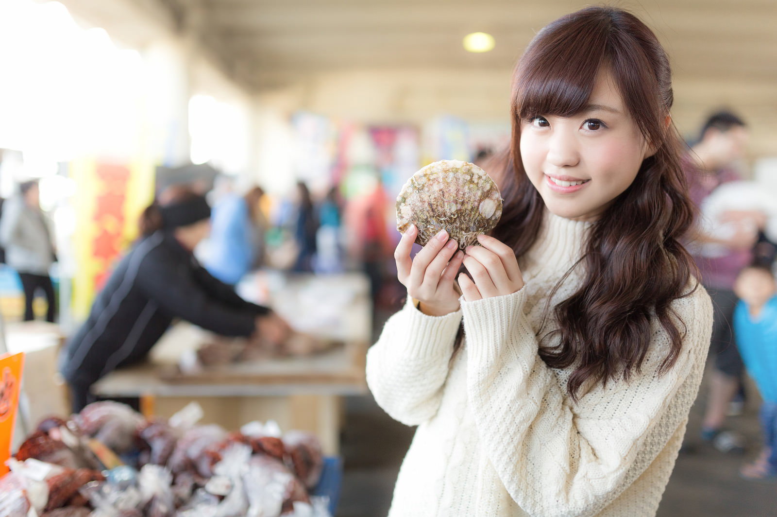 いすみ市の朝市で両手に持ったホタテをPRする女性［モデル：河村友歌］