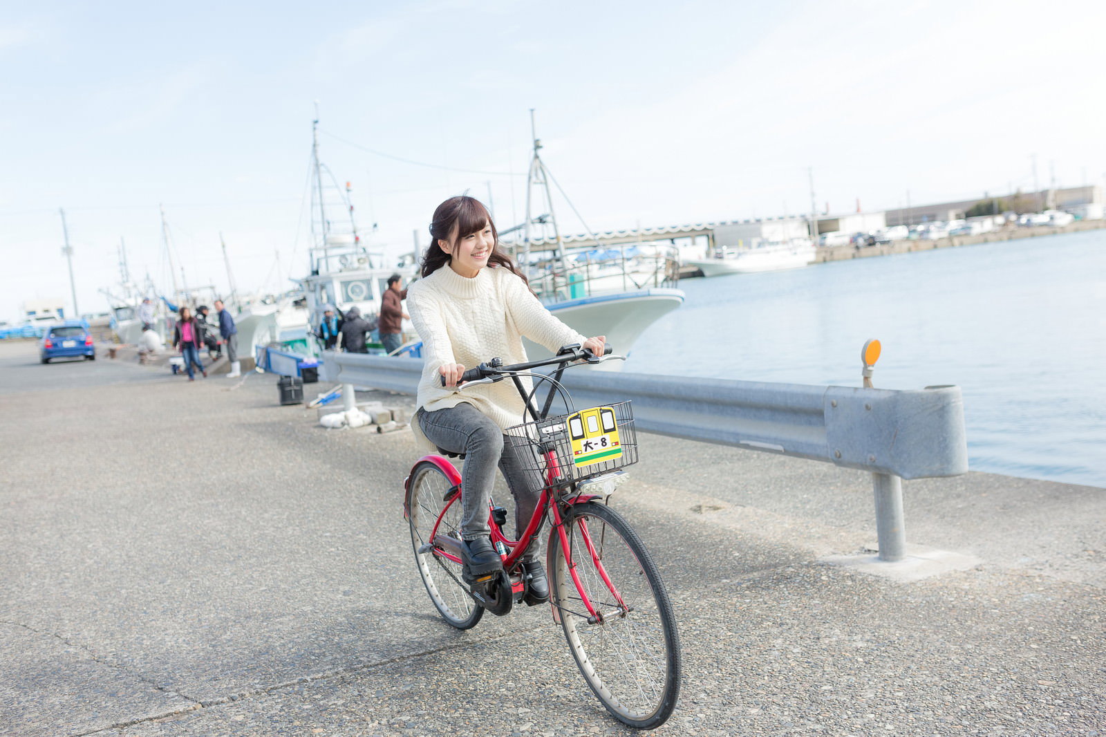 赤いレンタサイクルに乗って漁港沿いを走るクリーム色のセーターを着た女性［モデル：河村友歌］