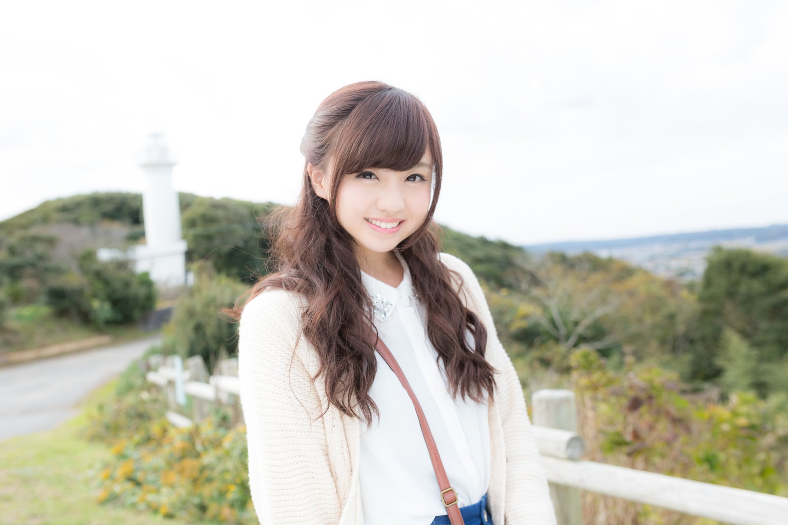 太東崎灯台を背景に笑顔で立つ女性。白いカーディガン姿でデート中［モデル：河村友歌］