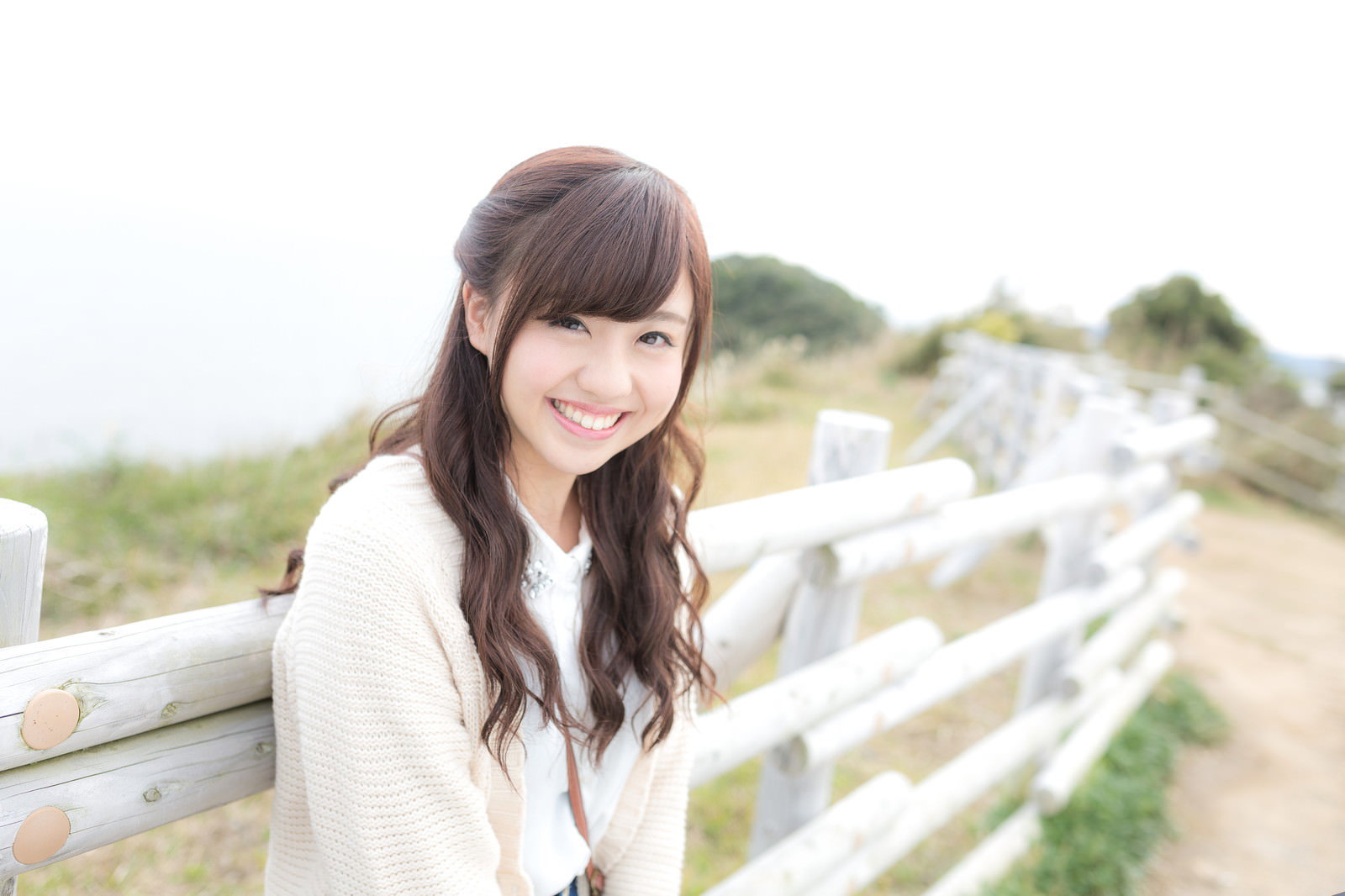 白いフェンスのそばに立ち笑顔を見せるクリーム色カーディガン姿の長髪女性［モデル：河村友歌］