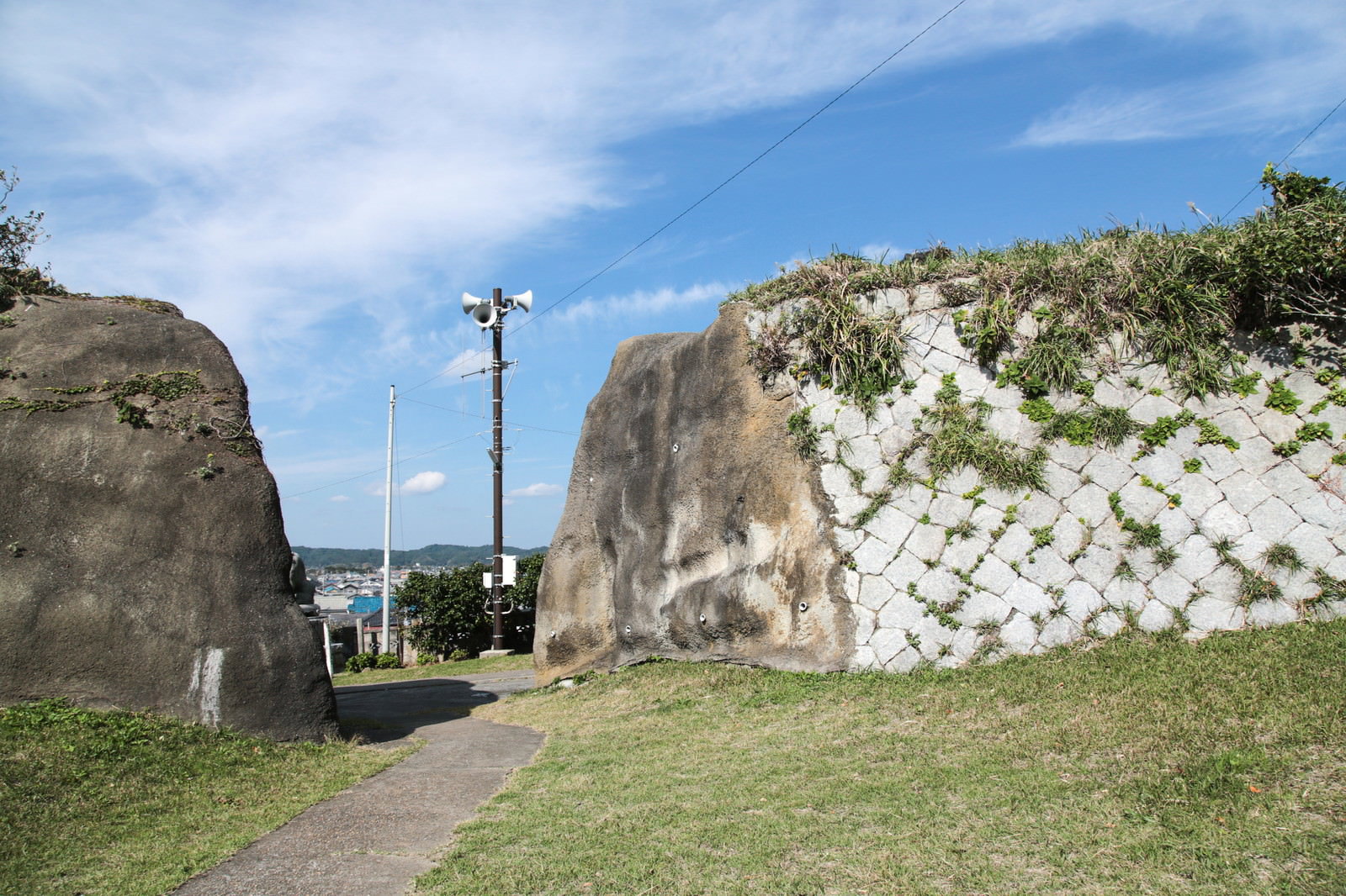 石積みの城跡に立つ電信柱と繁茂する緑、背景に海と町が見える景観