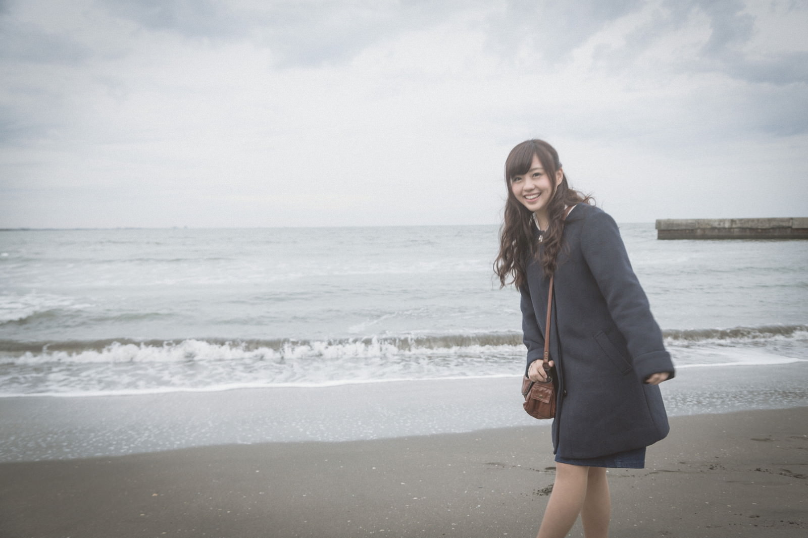 紺色のコートを着た女性が冬の海の波打ち際で笑顔を浮かべている［モデル：河村友歌］