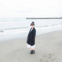 冬の海の波打ち際に立つお団子ヘアの女性、黒いコートで切ない表情の写真