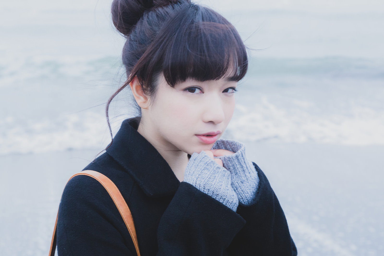 黒いコートとグレーのスカーフを身につけた女性が、冬の海岸で横向きに立ち、曇り空と波打ち際を背景に遠くを眺めている［モデル：渡辺友美子］