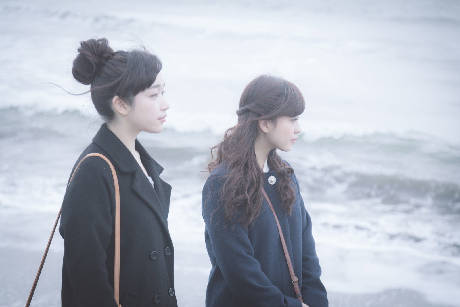 紺色のコートを着た二人の女性が秋冬の海辺に立ち、波を眺めている［モデル：河村友歌 渡辺友美子］