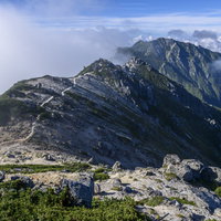 青空と雲が流れゆく南駒ヶ岳方面の登山道を行く登山者の写真
