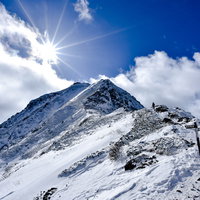 雪に覆われた赤岳山頂で光芒が差し込む中を歩む登山者の写真