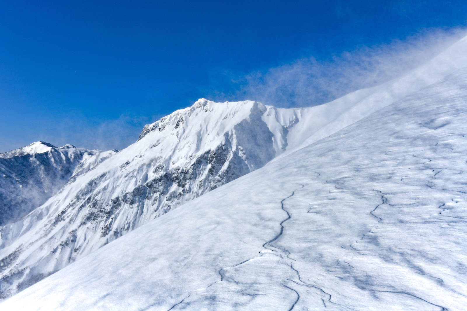 青空の下、雪に覆われた谷川岳中腹で地吹雪が舞い上がる冬山風景