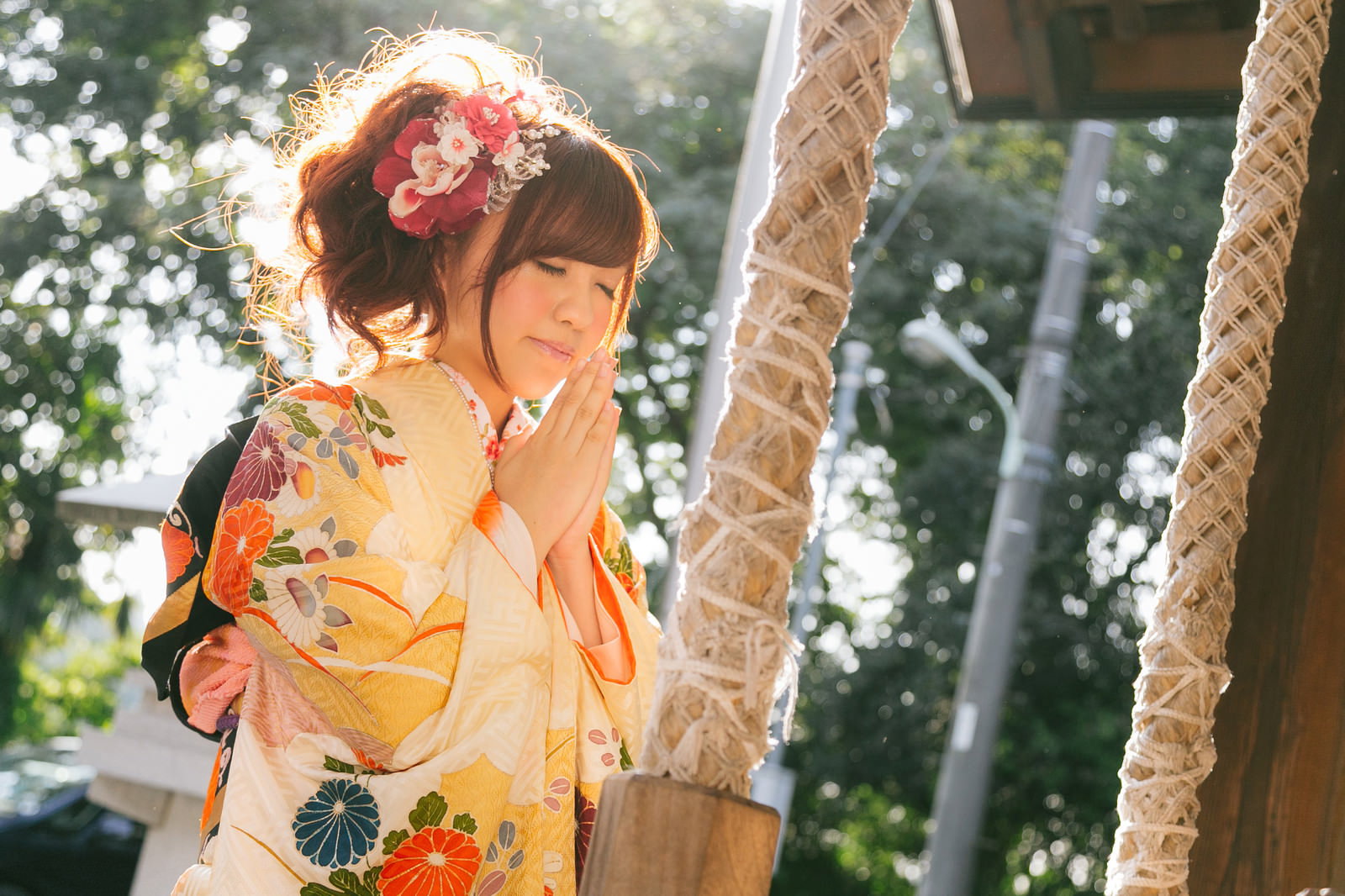 神社で鈴緒を握って参拝する赤い花飾りをつけた着物姿の女性［モデル：河村友歌］