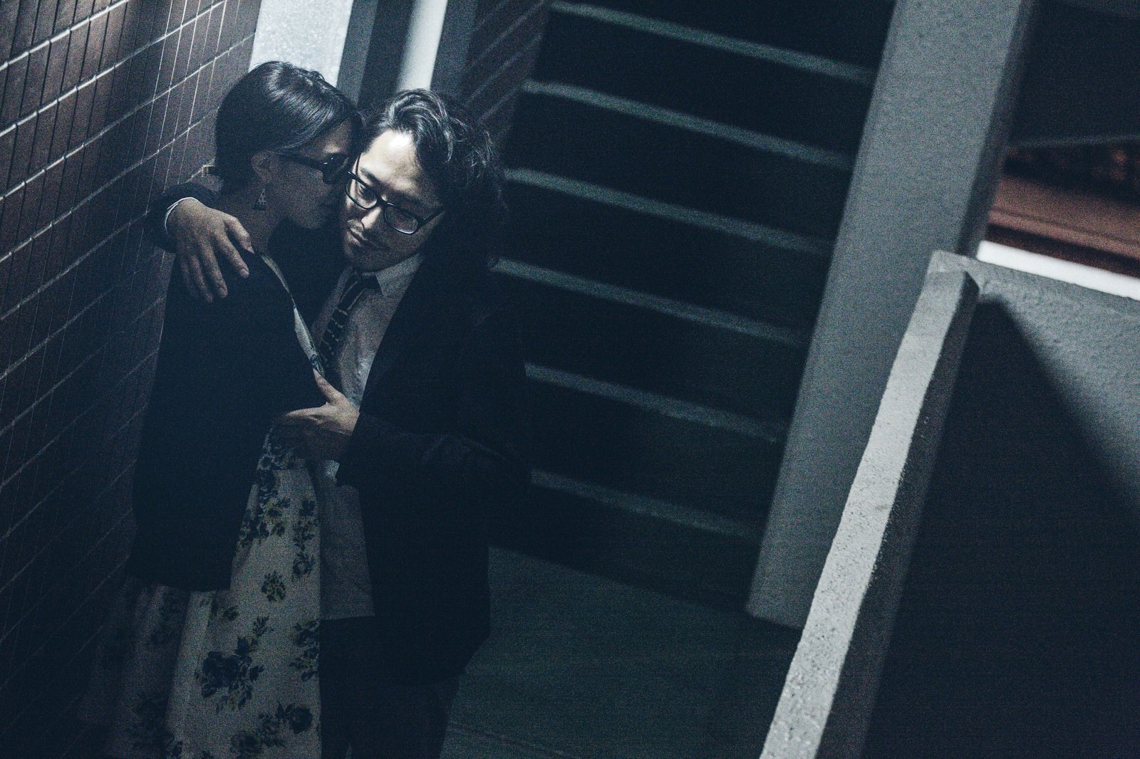 暗い階段で肩を寄せ合い密着する眼鏡の男性とスーツ姿の女性［モデル：ゆうせい］