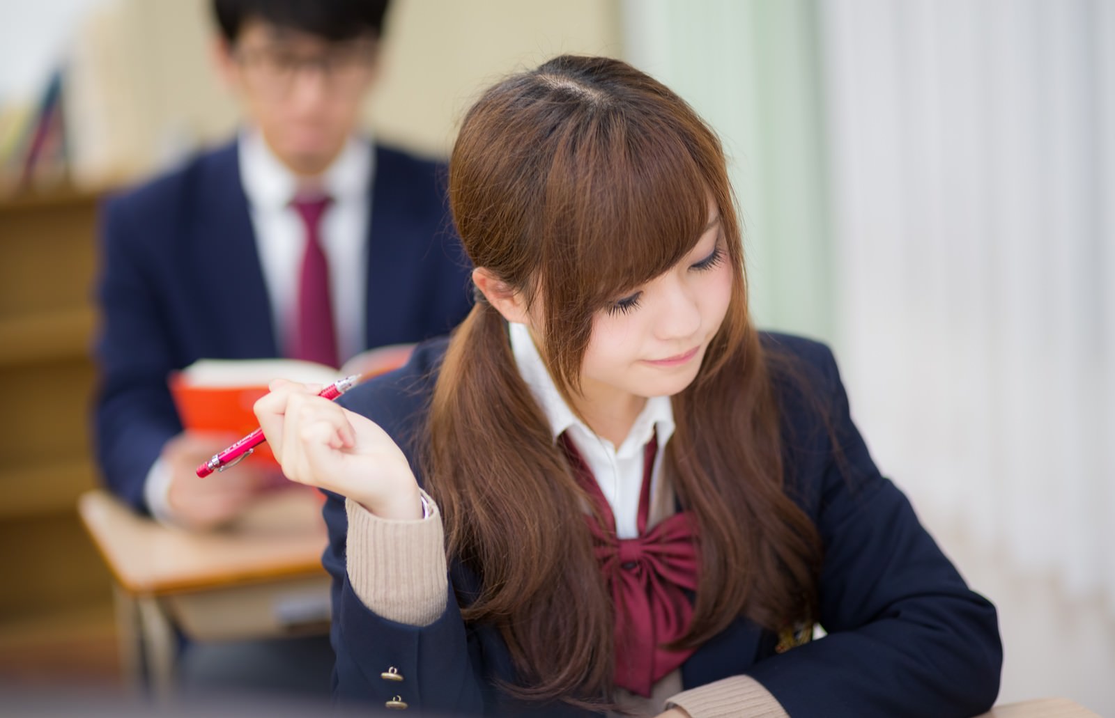 授業中に下を向いてペンを持つ制服姿の女子高生と背景の男子生徒［モデル：河村友歌 大川竜弥］