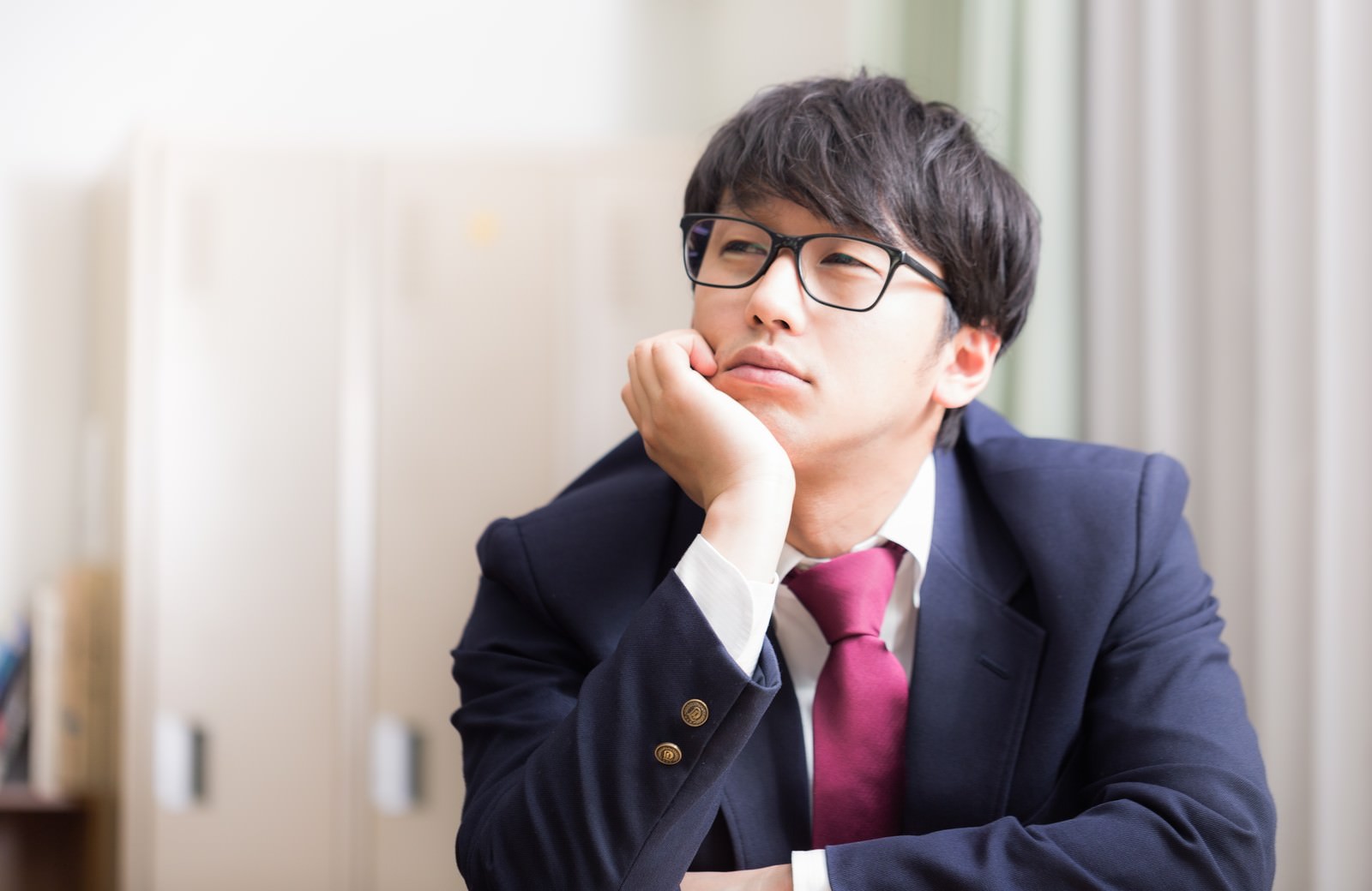 眼鏡をかけた男子高校生が学生服姿で頬杖をついて考え込んでいる［モデル：大川竜弥］