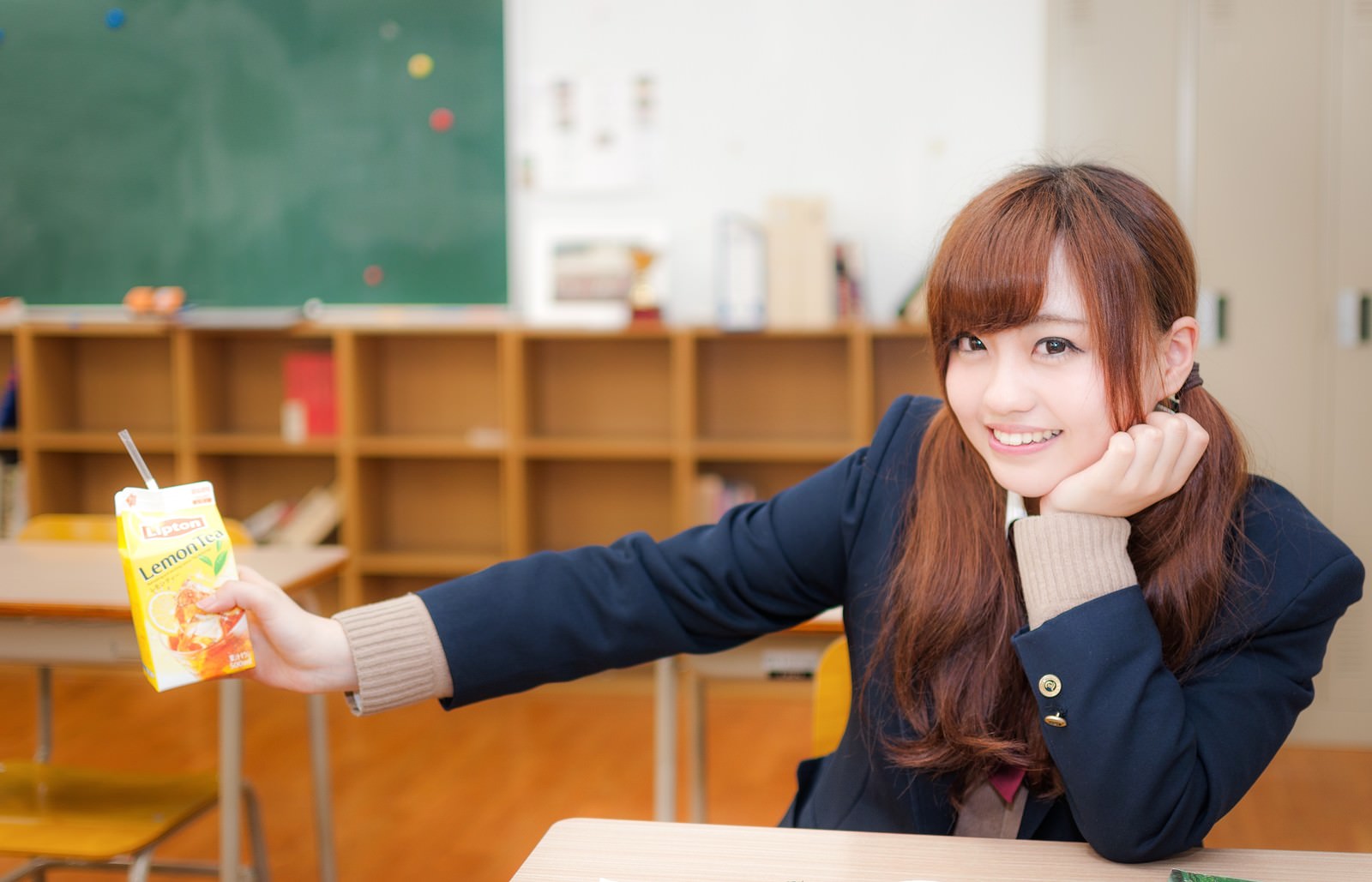 紺色の制服姿で教室の机に座り、レモンティーを持って笑う女子高生［モデル：河村友歌］
