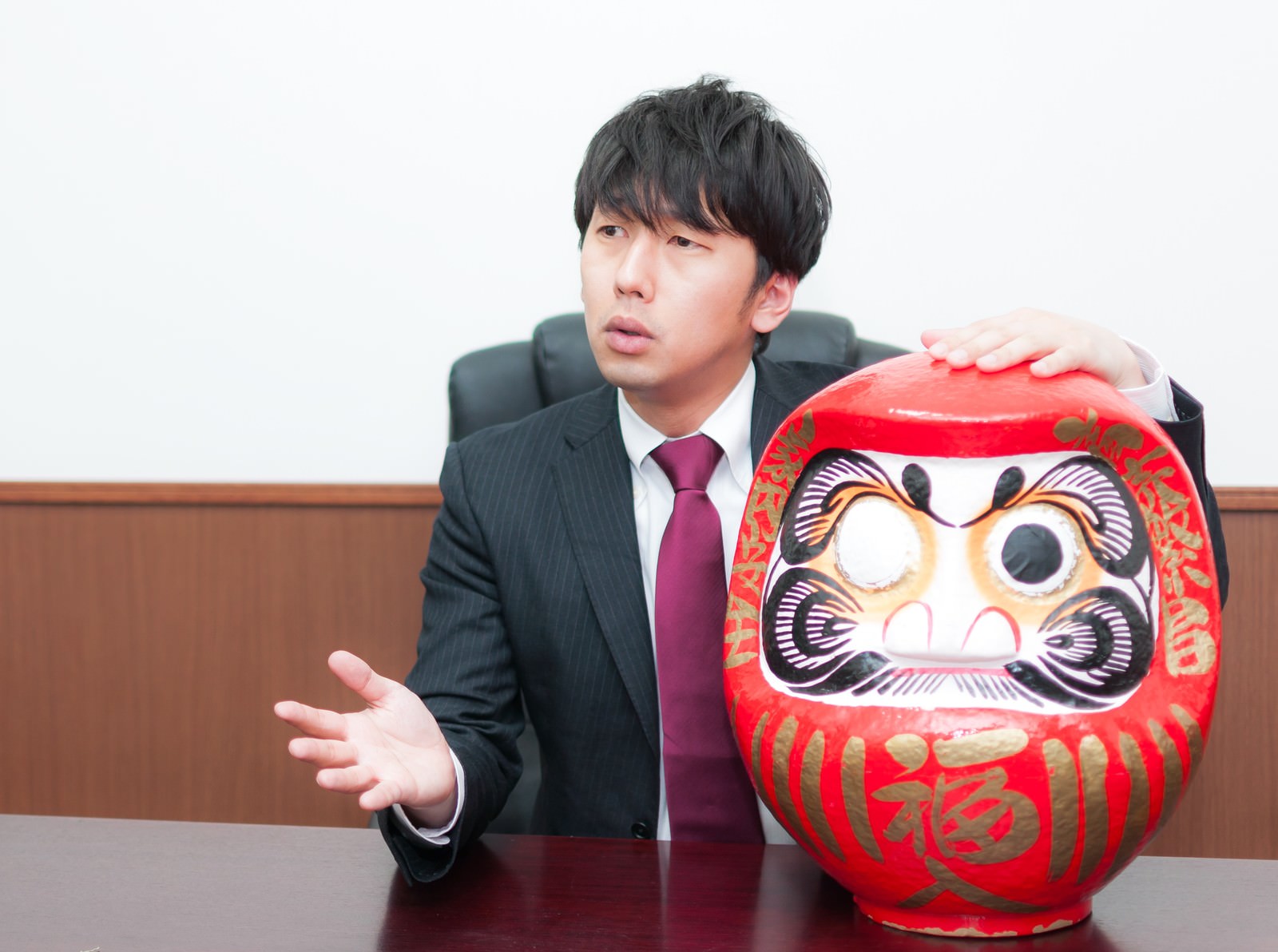 取材に応じる役員と片目の赤いだるま［モデル：大川竜弥］