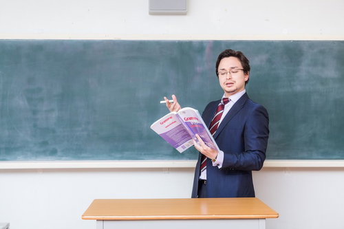 アメリカンジョークが通じないことに困惑する教師が教科書を手に黒板の前に立つ