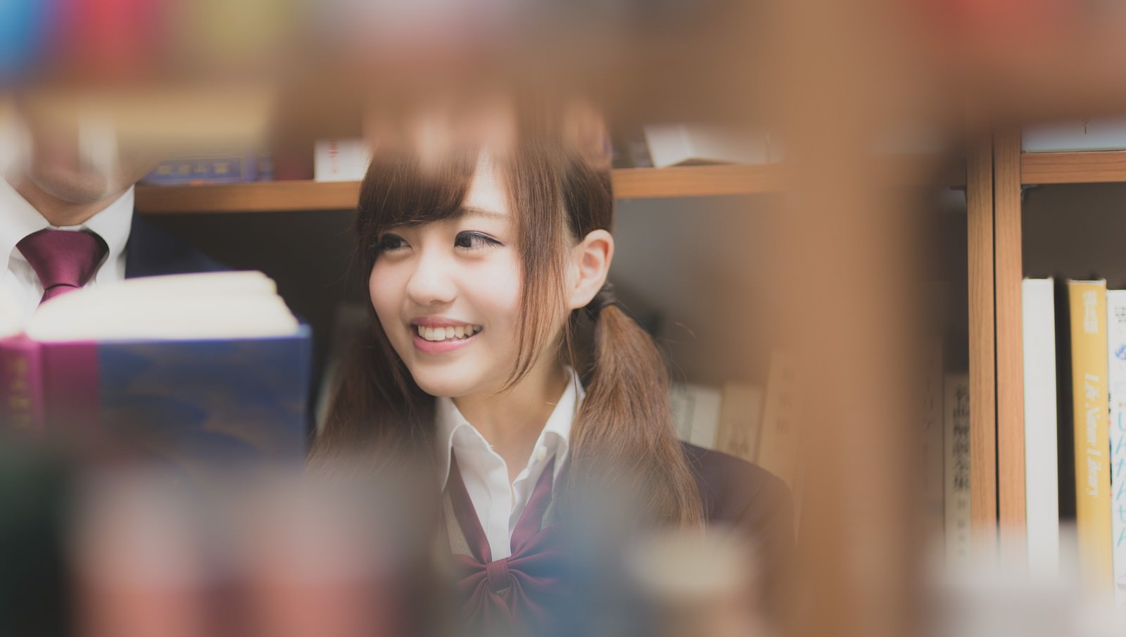 図書室で本棚越しに微笑む制服姿の女子高生［モデル：河村友歌］
