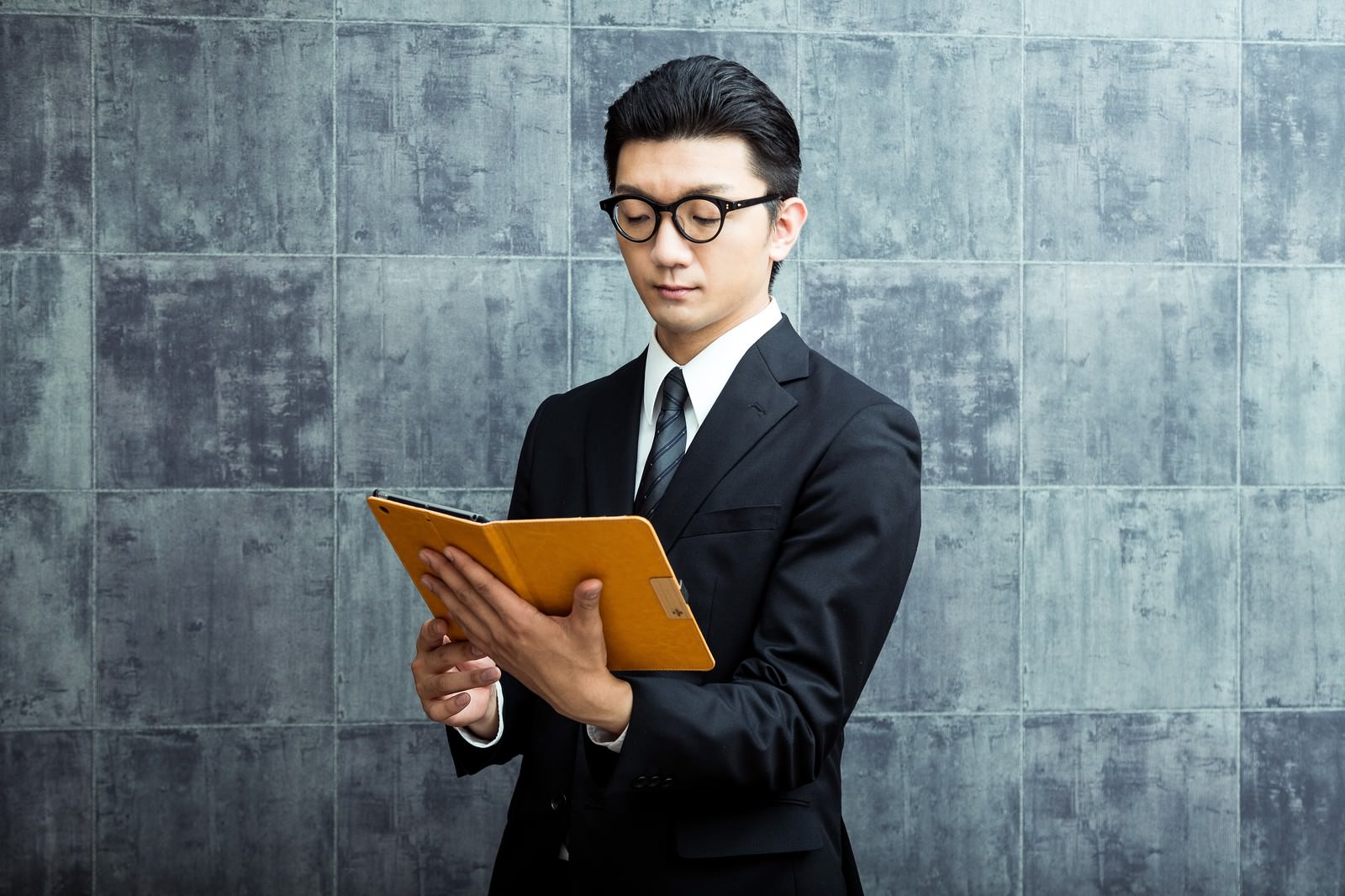 黒いスーツにメガネをかけた男性が、オレンジ色のタブレットで名簿を確認している［モデル：OZPA］