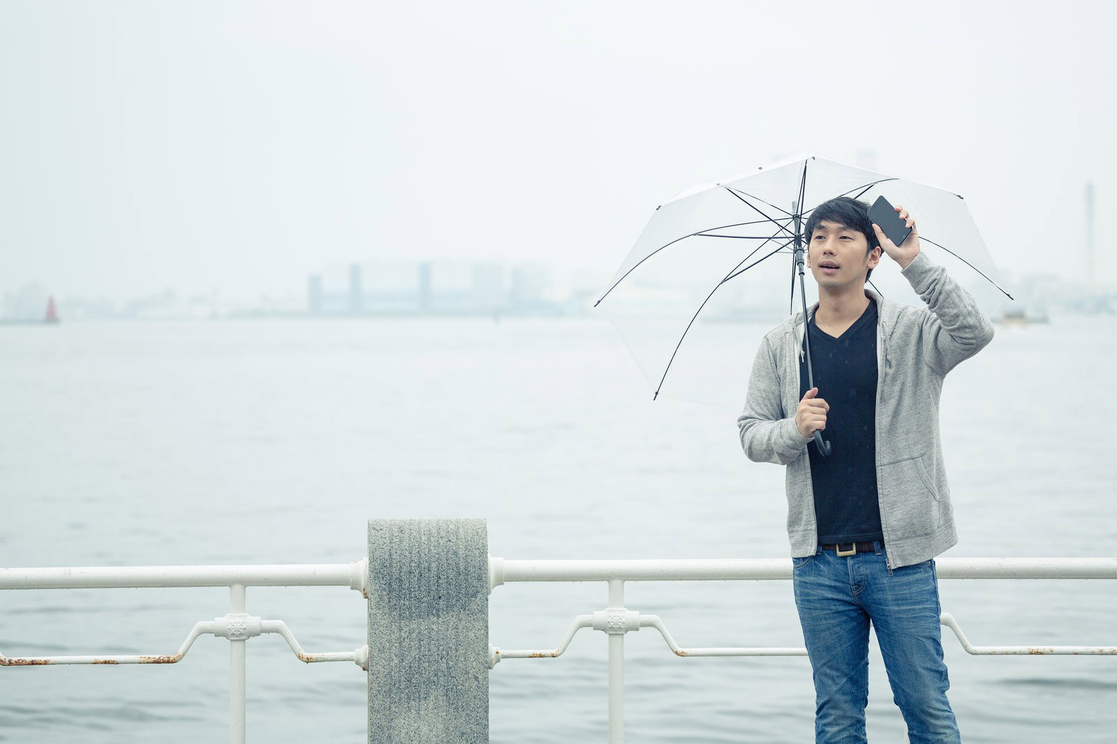 雨の中、スマートフォンを確認しながら傘をさす男性［モデル：大川竜弥］