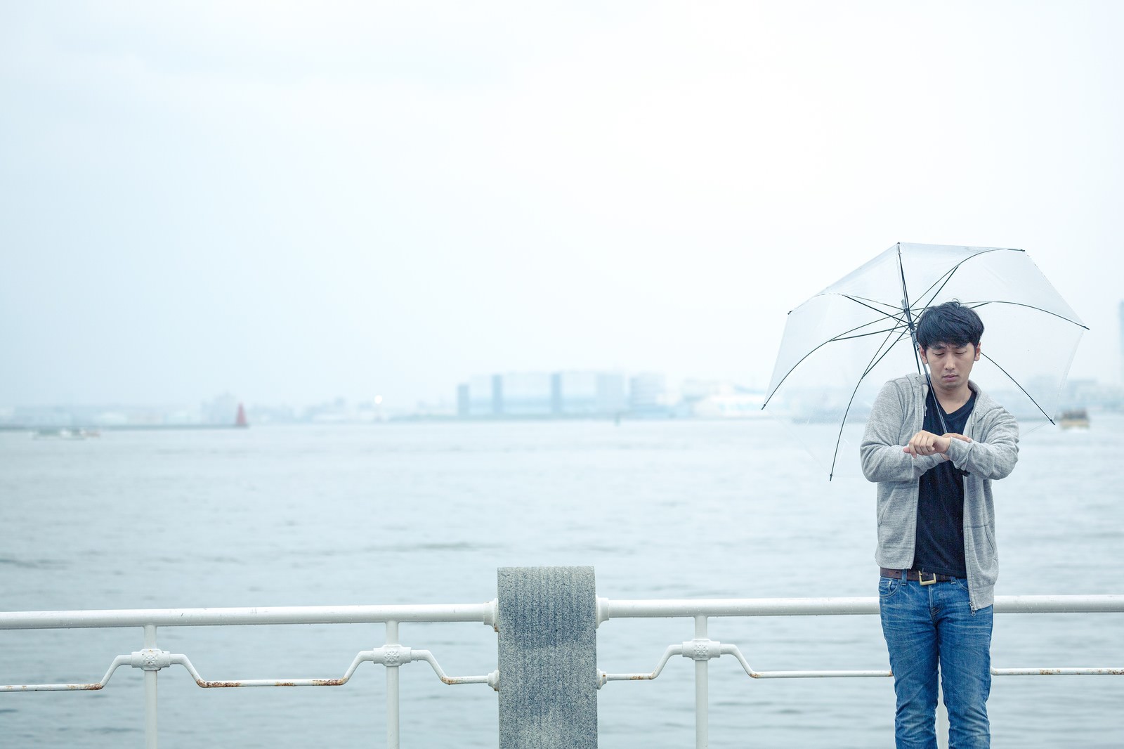 雨の中、埠頭で傘を持ちながら腕時計を確認する男性［モデル：大川竜弥］