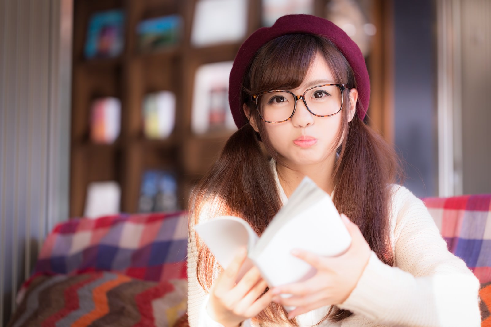 ワインレッド系ベレー帽とメガネをつけた女性が、クリーム色セーターで本を持って左上を見つめ、怒った表情で口を開けている［モデル：河村友歌］