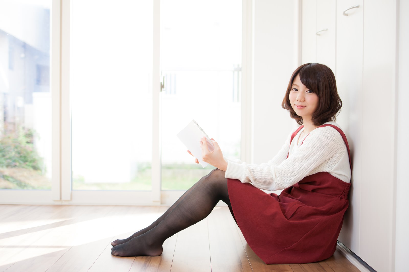 リビングでタブレットを持ち読書する女性が壁に寄りかかって座っている［モデル：みき。］