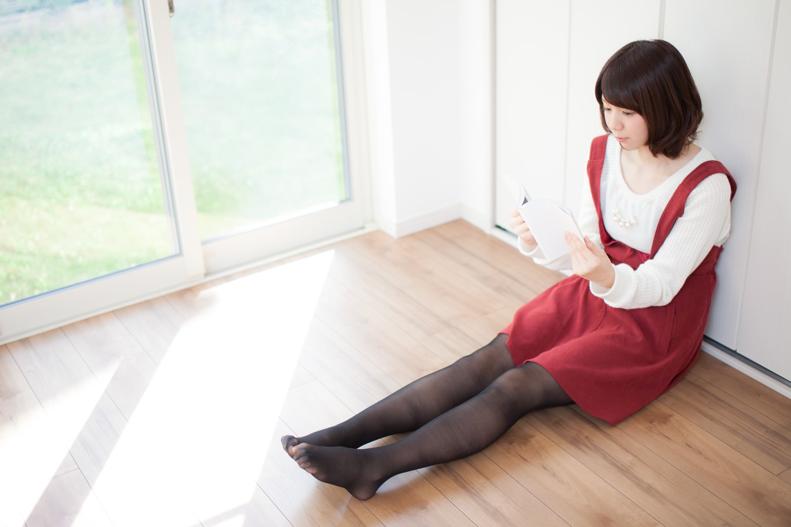 白い室内で床に座り、マンガ本を読む赤いベストの女性。窓からの光が当たるリビング空間。［モデル：みき。］
