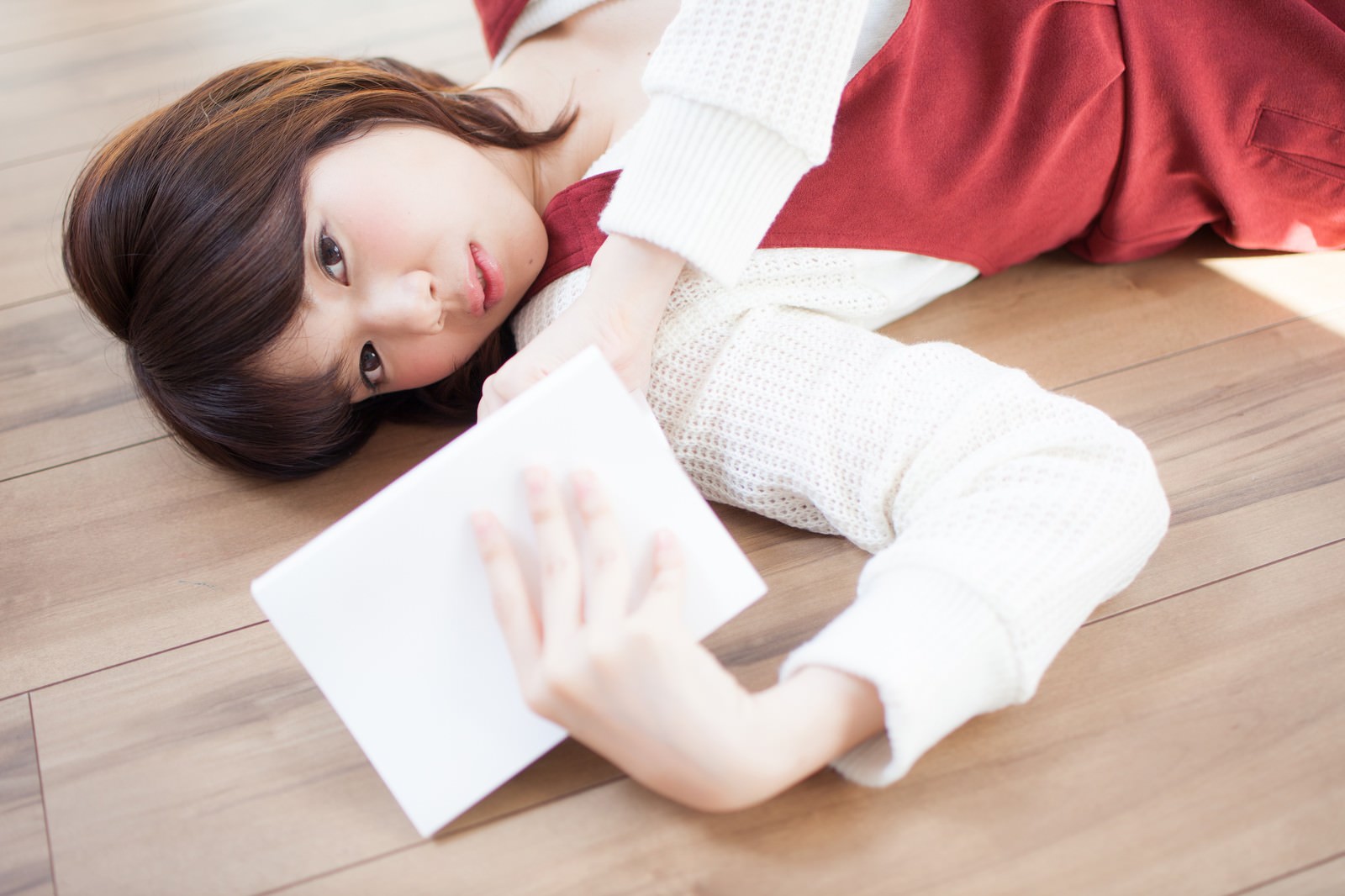 白いニットと赤いスカート姿の女性がフローリングに寝転び本を読んでいる［モデル：みき。］