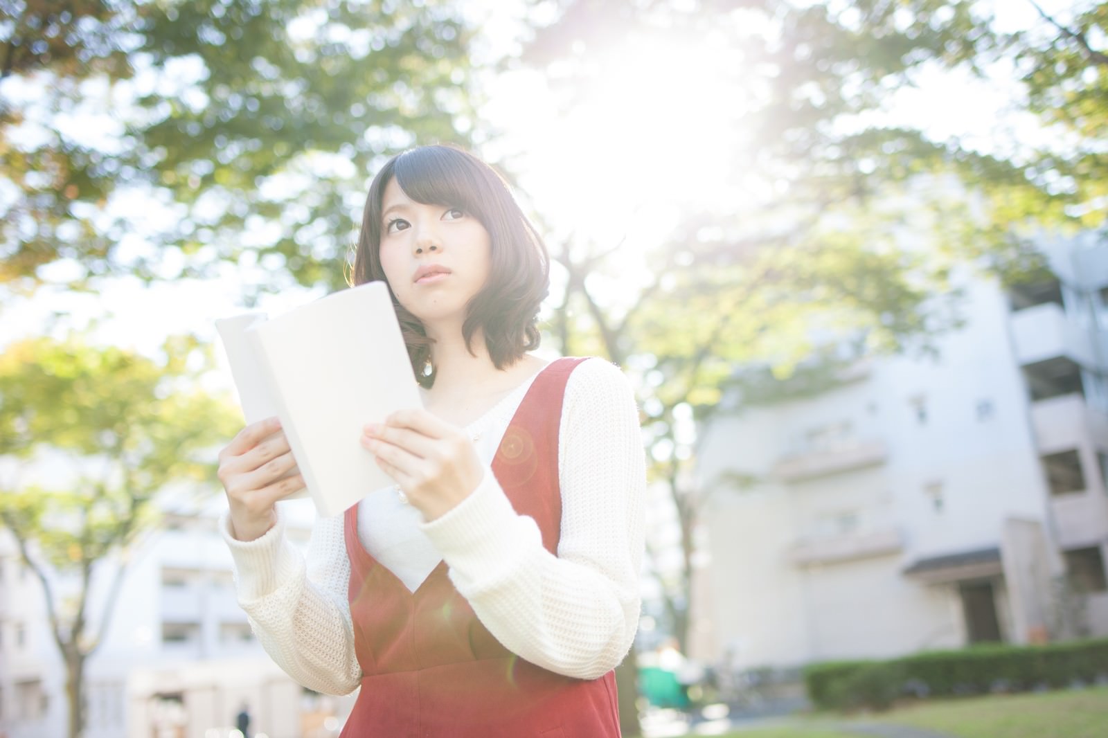緑の樹木の下で本を読む赤いベストの女性［モデル：みき。］