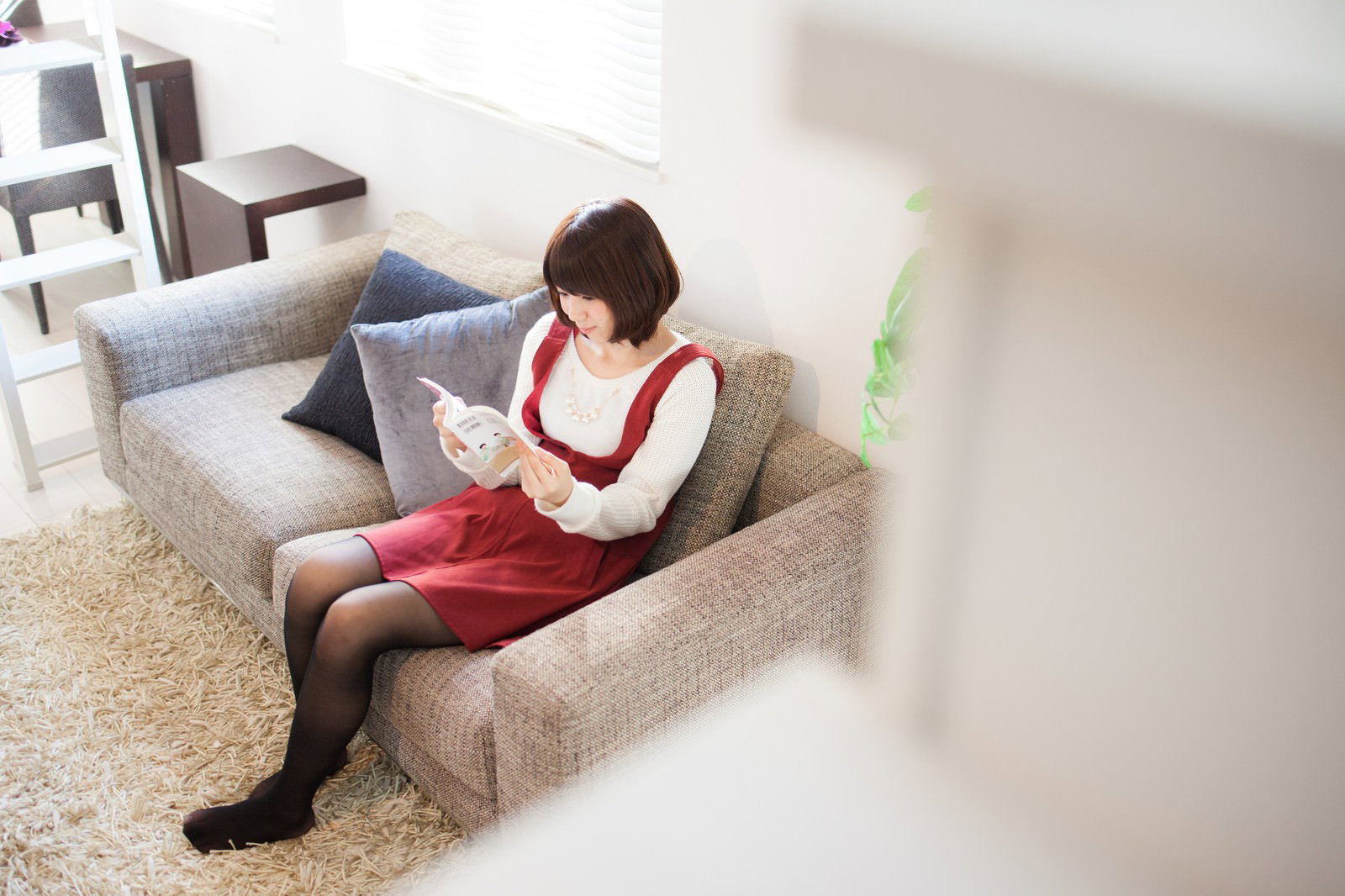 ソファーでマンガを読む赤いベストの女性［モデル：みき。］