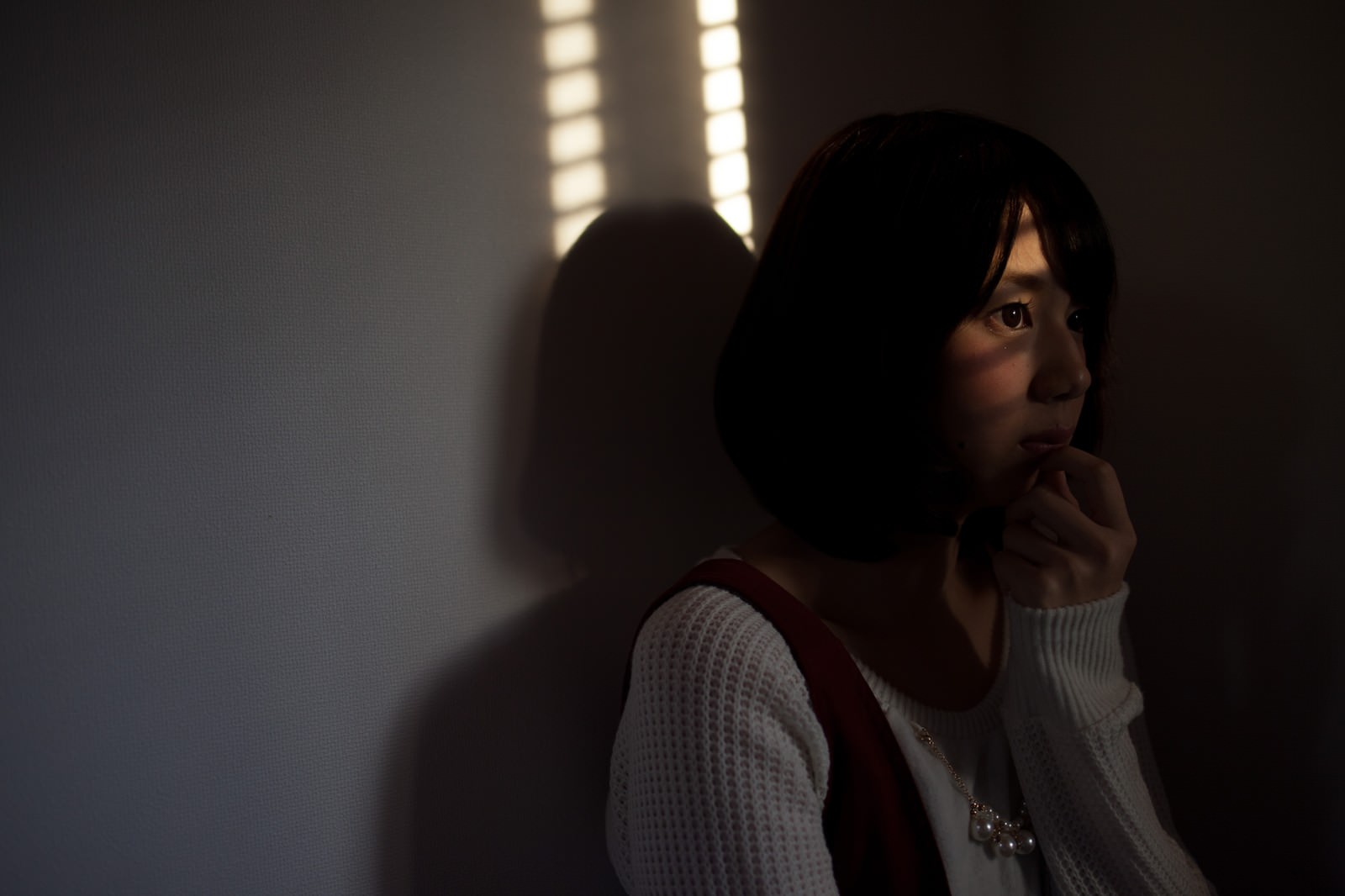 ブラインドごしに外を見つめ、手を口に当てて驚く女性。背後に人物の影がぼやけて映る［モデル：みき。］