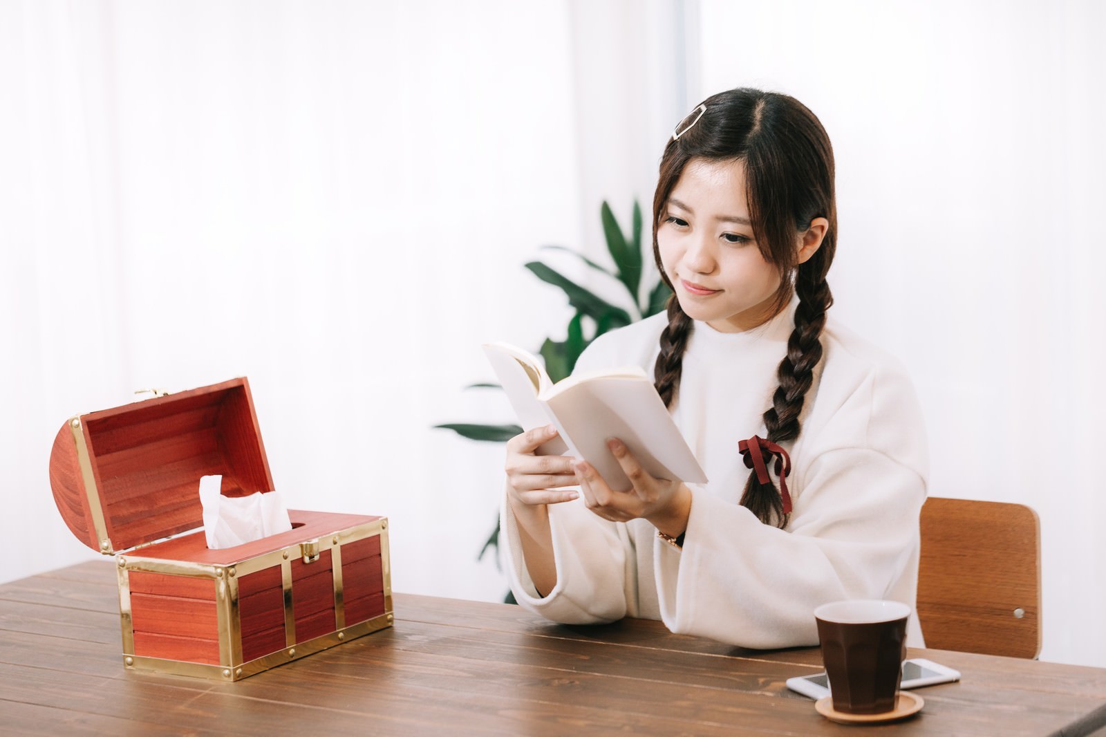 白いシャツを着た三つ編みの女性が、木製テーブルで本を読んでいる。奥に赤い宝箱のティッシュボックス［モデル：河村友歌］