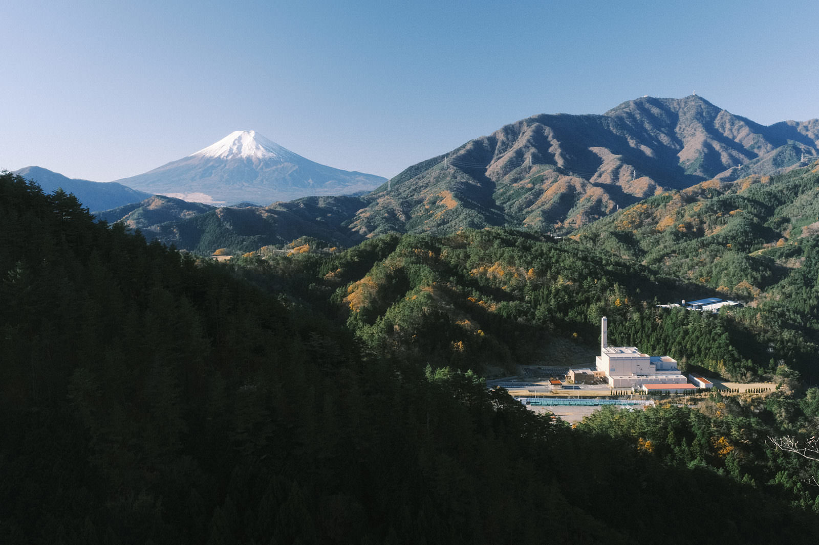 青空の下、緑の山間に富士山がそびえ立ち、手前に白い建物がある風景