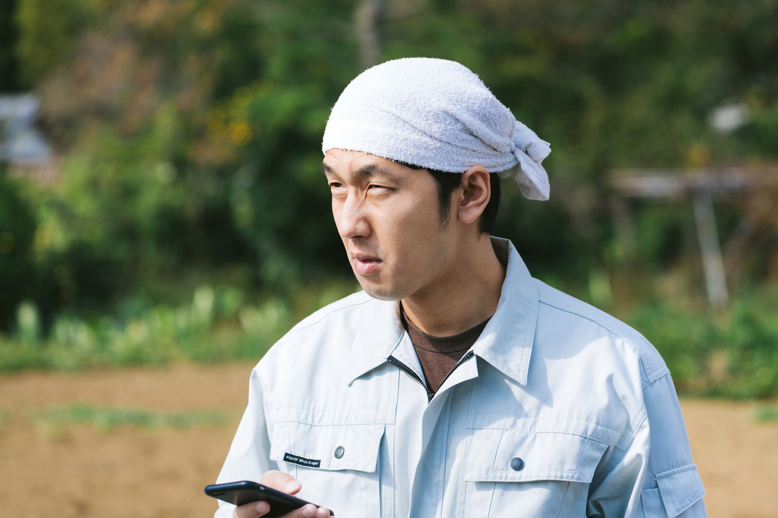 白いタオルを頭に巻いた農民の男性がスマートフォンを持ち、困った表情で横を向いている［モデル：大川竜弥］