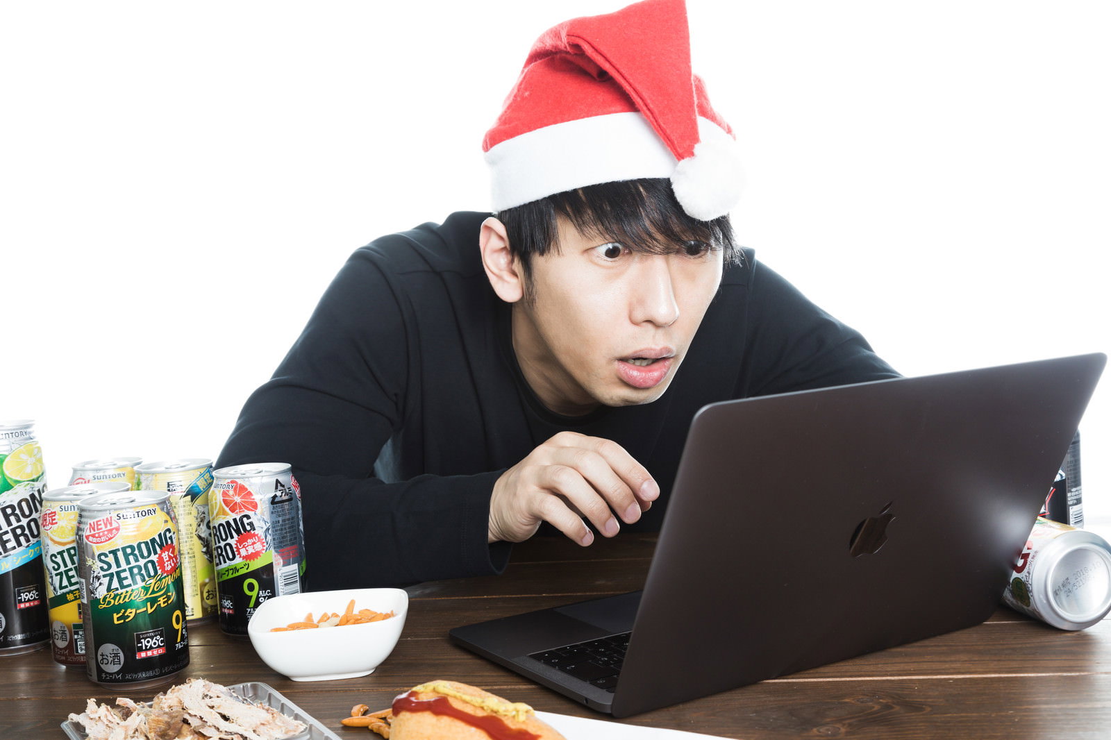 サンタ帽を被ってノートパソコンでバグ対応に驚く男性、缶飲料とスナック菓子に囲まれたデスク［モデル：大川竜弥］