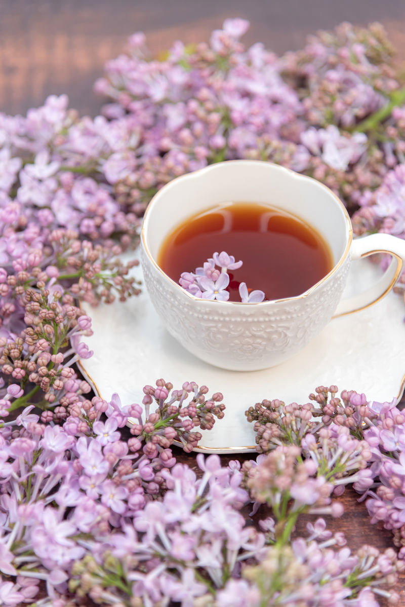 薄紫色のライラックの花に囲まれた白いティーカップと琥珀色の紅茶