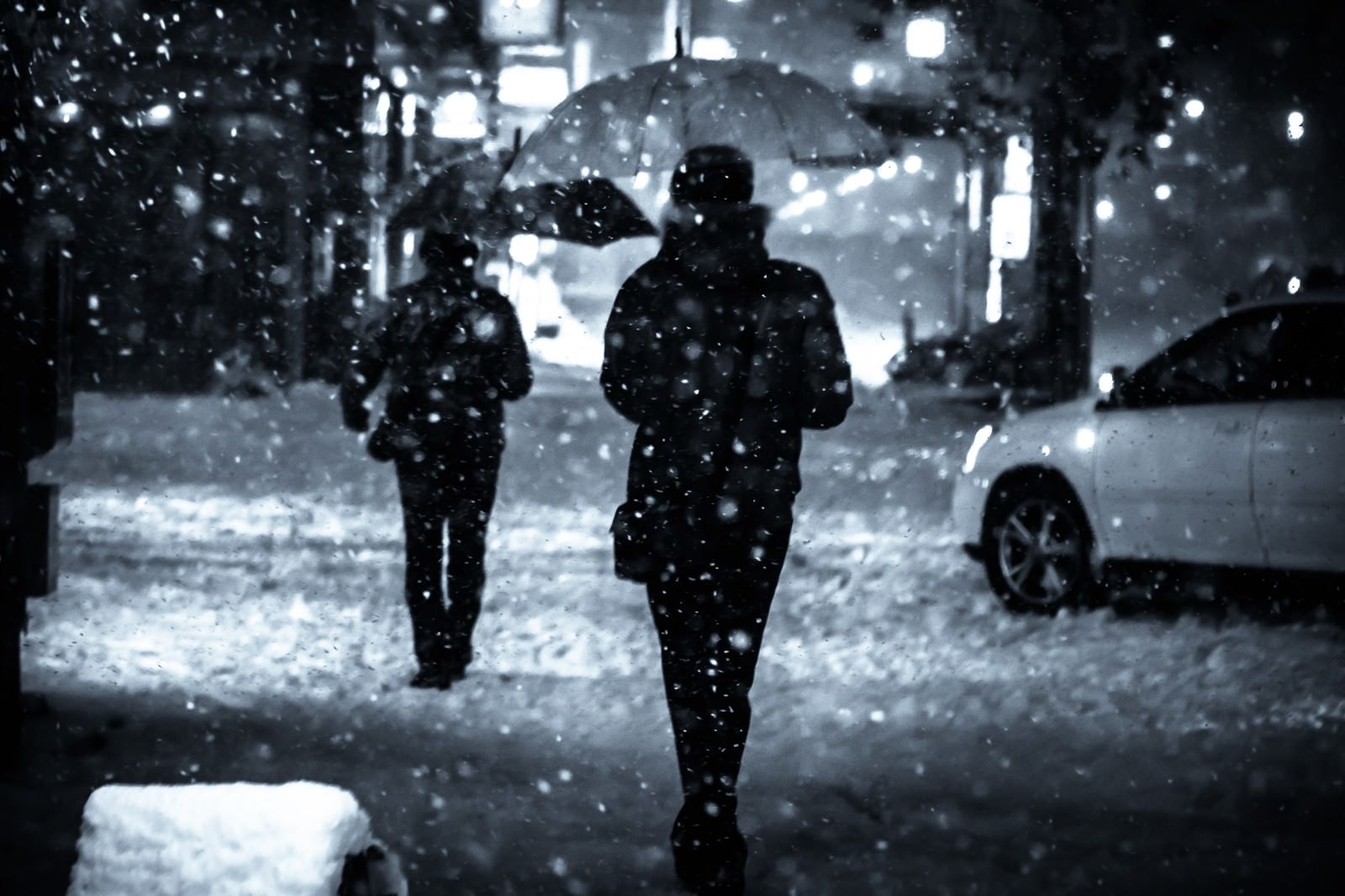 夜間の雪が降る街で傘をさして歩く人物たちのシルエット