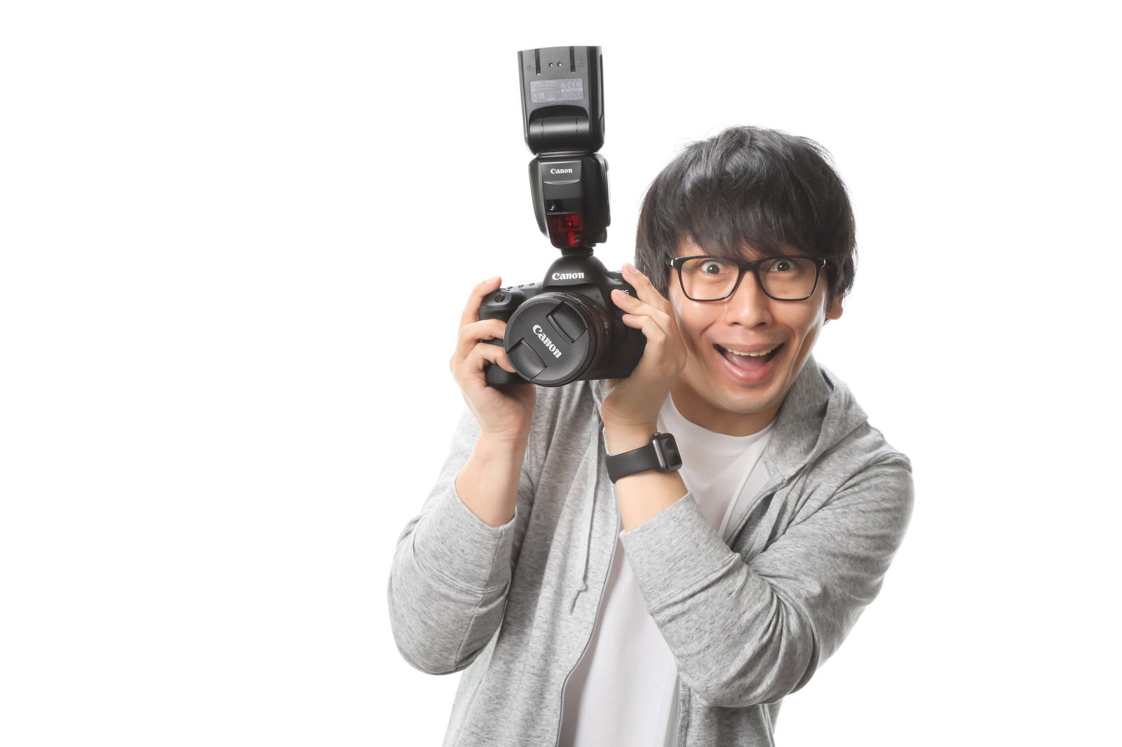 Canon一眼レフカメラとストロボを構えて笑顔で見つめる眼鏡男性［モデル：大川竜弥］