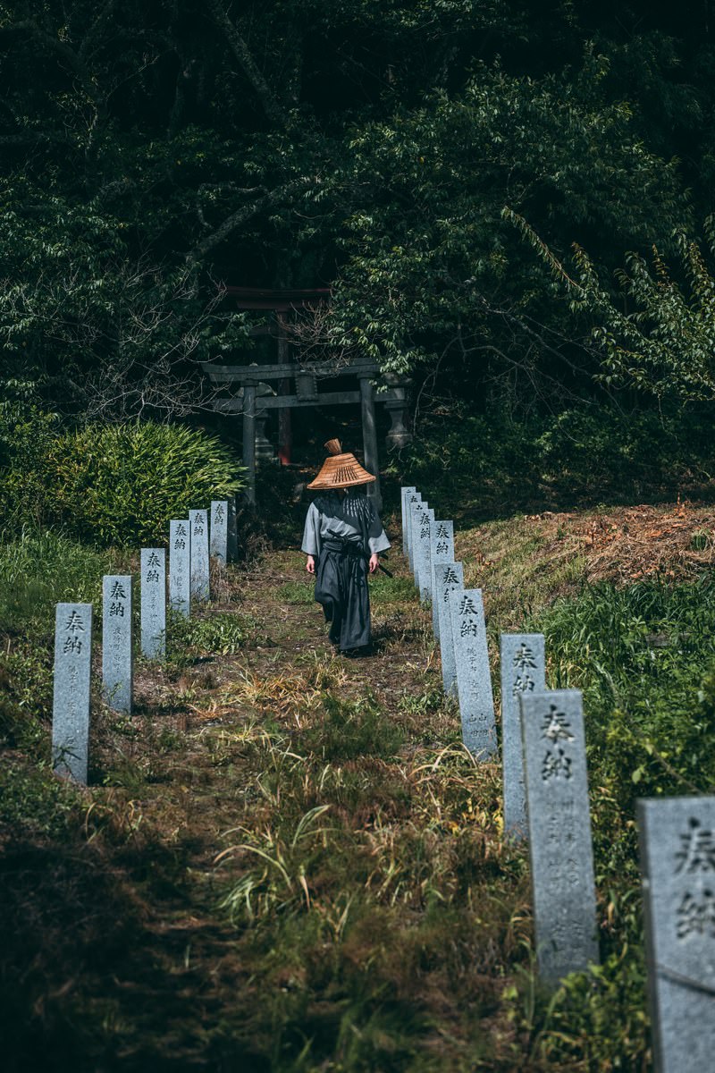 奉納石柱が並ぶ参道を歩き、鳥居へ向かう笠をかぶった侍の後ろ姿［モデル：まーこ］