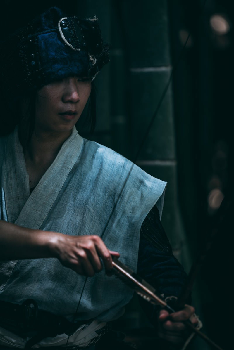 大弓を構える甲冑装備の野盗男性が薄紫の衣装で伏し目がちに立つ［モデル：まーこ］