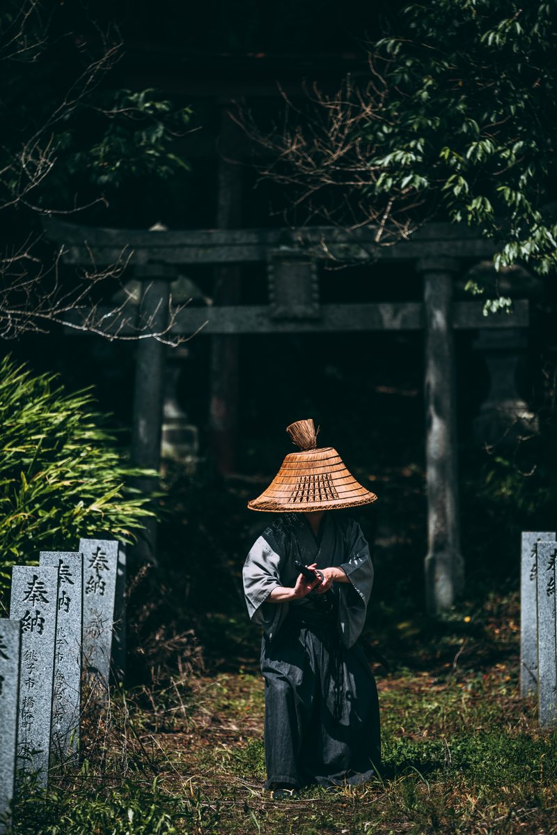 茶色い笠をかぶり、黒い和装で刀に手をかけた男性が、神社の鳥居前の奉納石柱の側に立つ［モデル：まーこ］
