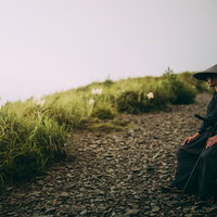 登山道脇の岩に腰掛けて休憩する笠かぶりの侍の写真
