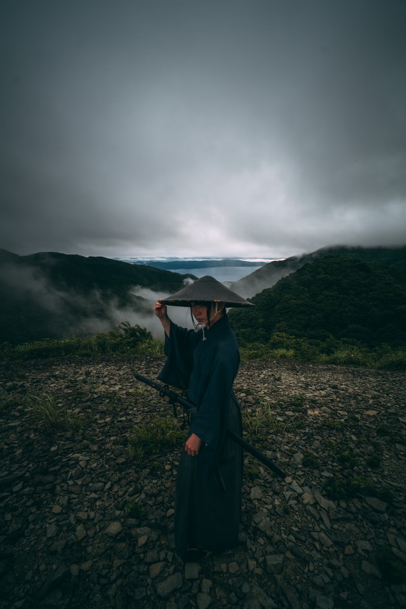 霧の中で笠を掲げる侍姿の男性。背後に湖と曇り空が広がる山景色［モデル：まーこ］